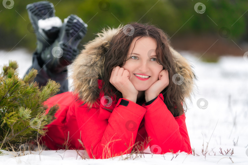 Скачать Симпатичная брюнетка с длинными вьющимися волосами лежит на зимнем снегу в хвойном лесу, улыбается и смотрит в сторону фотосток Ozero