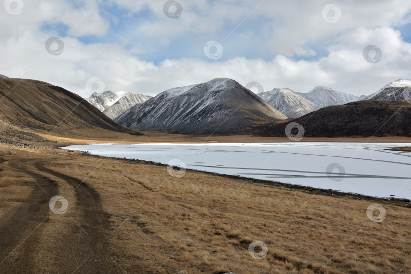 Скачать Солнечным осенним днем грязная полевая дорога вьется вдоль берега широкого замерзшего озера у подножия заснеженных гор. фотосток Ozero