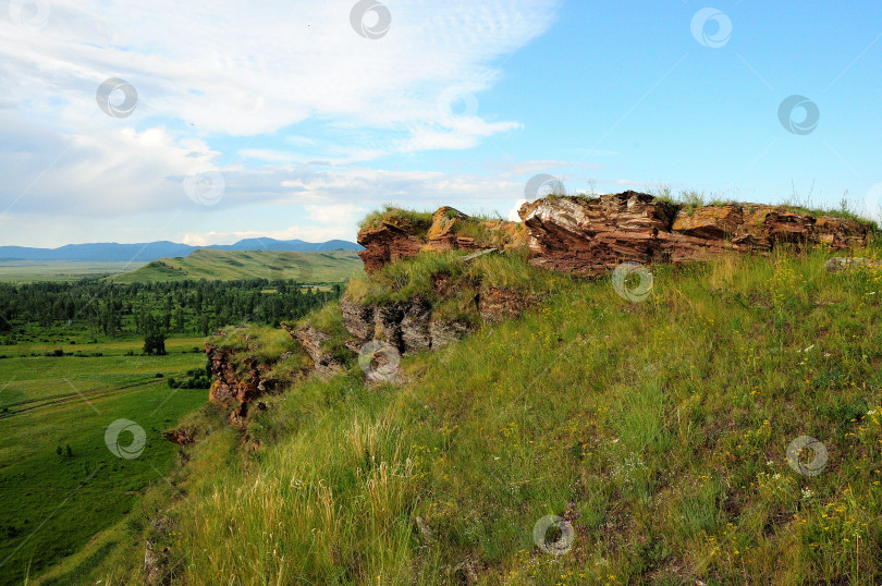 Скачать Слоистые скальные образования из красного песчаника на вершине высокого холма с видом на обширную летнюю долину. фотосток Ozero
