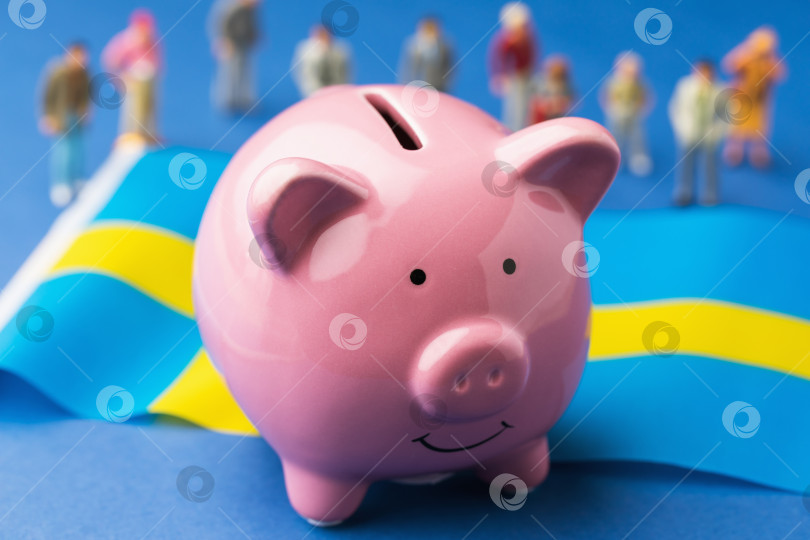Скачать Керамическая копилка, шведский флаг и пластиковые игрушечные человечки на синем фоне, концепция на тему доходов населения Швеции фотосток Ozero