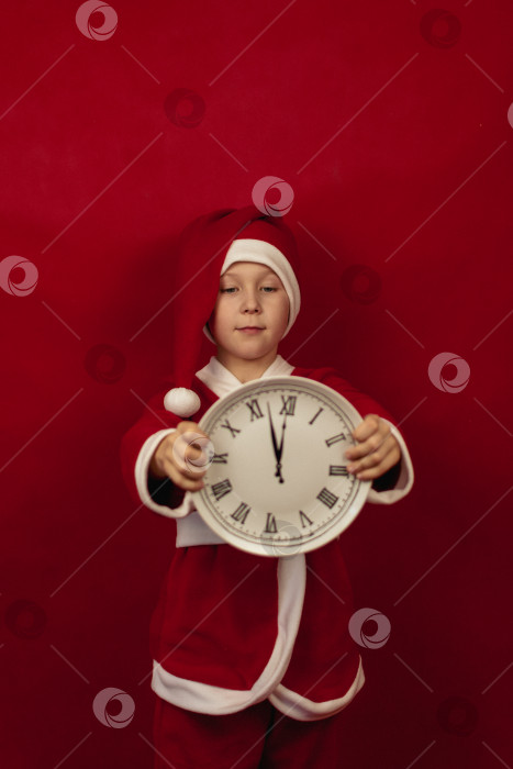 Скачать Мальчик в костюме Санты держит в руках белые часы на красном фоне. фотосток Ozero