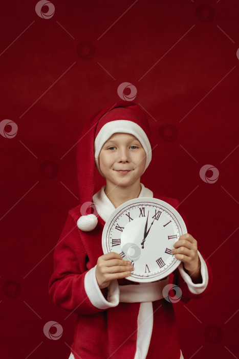 Скачать Мальчик в костюме Санты держит в руках белые часы на красном фоне. фотосток Ozero