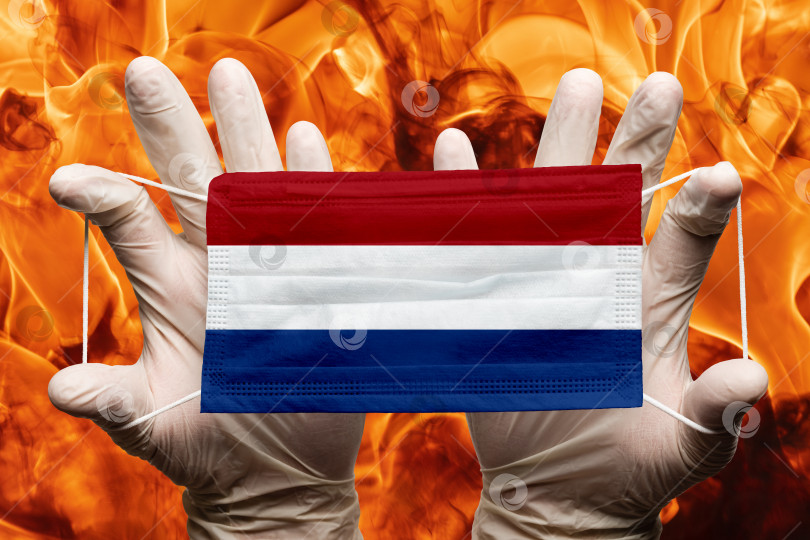 Скачать Врач держит в белых перчатках защитную медицинскую маску для лица, респираторную повязку с наложенным на маску национальным флагом Нидерландов. Концепция фотосток Ozero