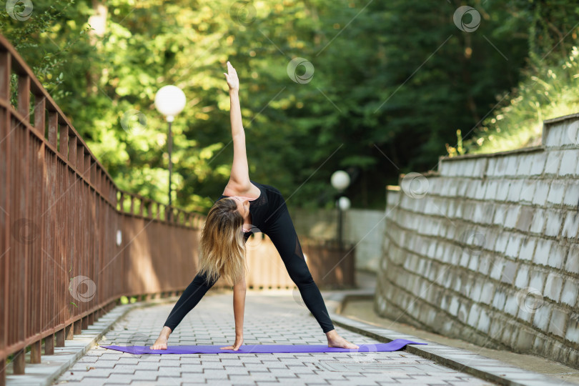 Скачать Женщина, практикующая йогу, выполняющая скручивающее упражнение Паривритта Падоттанасана, тренирующаяся летним утром в парке, стоя на коврике фотосток Ozero
