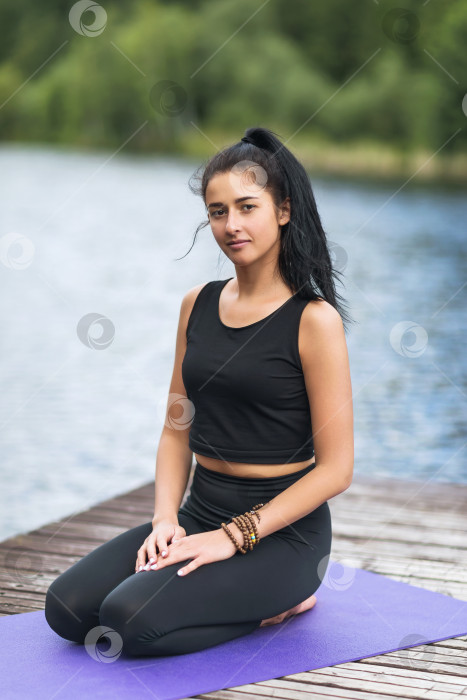 Скачать Портрет молодой красивой женщины-тренера по йоге, сидящей на деревянном пешеходном мостике на берегу озера фотосток Ozero