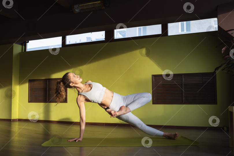 Скачать Молодая девушка, ведущая здоровый образ жизни и практикующая йогу, выполняет вариацию упражнения васиштхасана, позу мудреца, тренируется одна в белой спортивной одежде в комнате на коврике фотосток Ozero