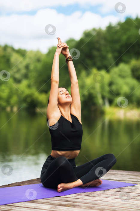 Скачать Девушка на деревянном мосту у реки, практикующая асаны йоги, выполняющая позу лотоса с поднятыми вверх руками фотосток Ozero