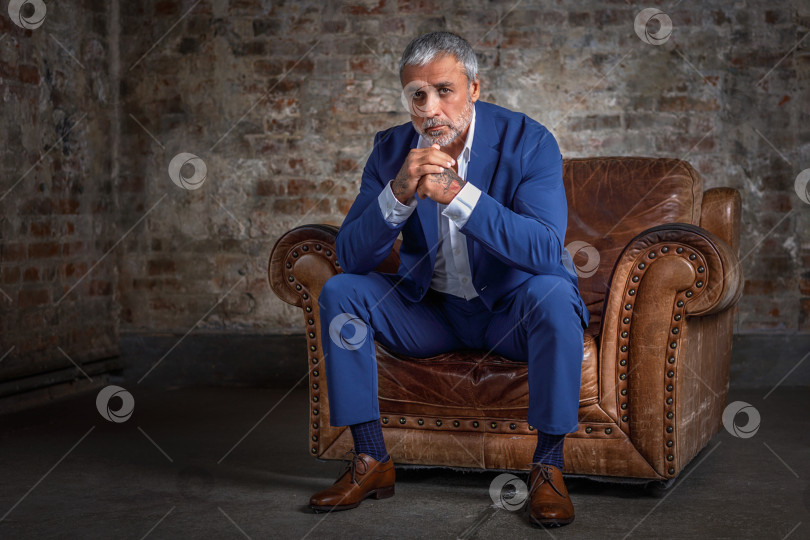 Скачать Красивый мужчина средних лет в синем строгом костюме позирует, сидя в кожаном кресле. Портрет бизнесмена. Красивый мужчина средних лет позирует на кирпичном фоне с местом для копирования фотосток Ozero