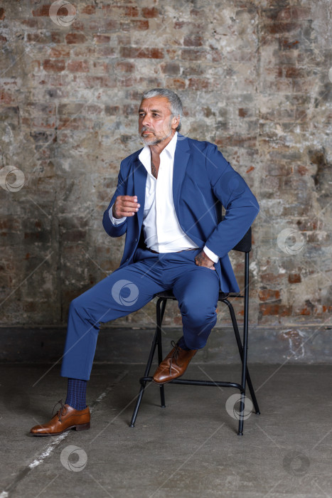 Скачать Красивый мужчина средних лет в синем строгом костюме позирует, сидя в кресле. Портрет бизнесмена. Красивый мужчина средних лет позирует на кирпичном фоне с местом для копирования фотосток Ozero