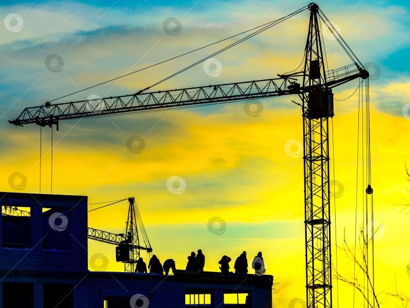 Скачать Силуэт бетонного каркаса строительной площадки высотного жилого дома в городе, строители, строительный кран, освещенный подсветкой солнечного света заката или рассвета. фотосток Ozero