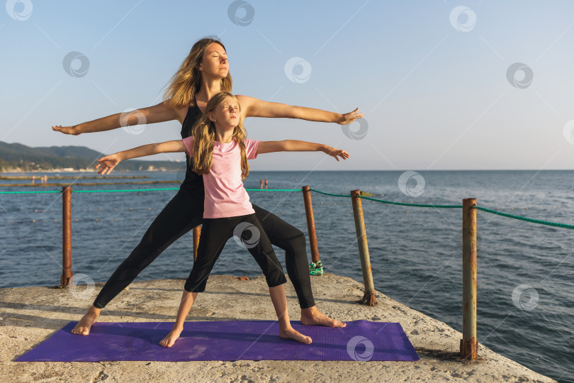 Скачать Мама и дочь, практикующие йогу, солнечным летним вечером на пирсе у моря, стоя на коврике, выполняют упражнение вирабхадрасана, поза воина фотосток Ozero