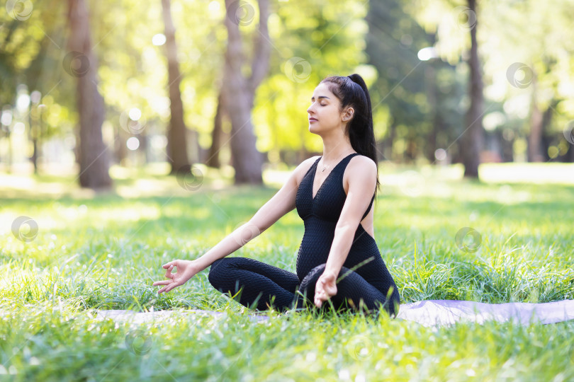 Скачать Молодая беременная девушка в черном спортивном комбинезоне, практикующая йогу, выполняет медитацию, сидя в позе лотоса на коврике в парке фотосток Ozero