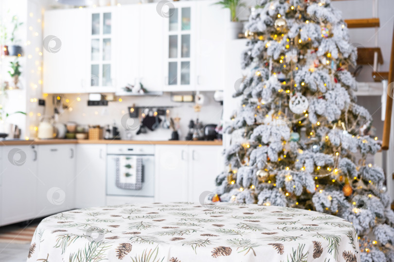 Скачать Круглый пустой стол для демонстрации продукта со скатертью с принтом из сосновых иголок и шишек, место для текста на белой кухне, современный интерьер с рождественской елкой. Новый год, Рождество фотосток Ozero