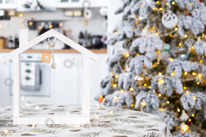 Скачать Ключ и крошечный домик небольшого размера в уютном доме с рождественским декором на столе праздничной белой кухни. Подарок на Новый год. Страхование, переезд в новый дом, ипотека, аренда и покупка недвижимости фотосток Ozero