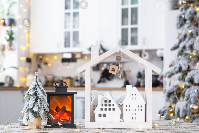 Скачать Ключ и крошечный домик небольшого размера в уютном доме с рождественским декором на столе праздничной белой кухни. Подарок на Новый год. Страхование, переезд в новый дом, ипотека, аренда и покупка недвижимости фотосток Ozero