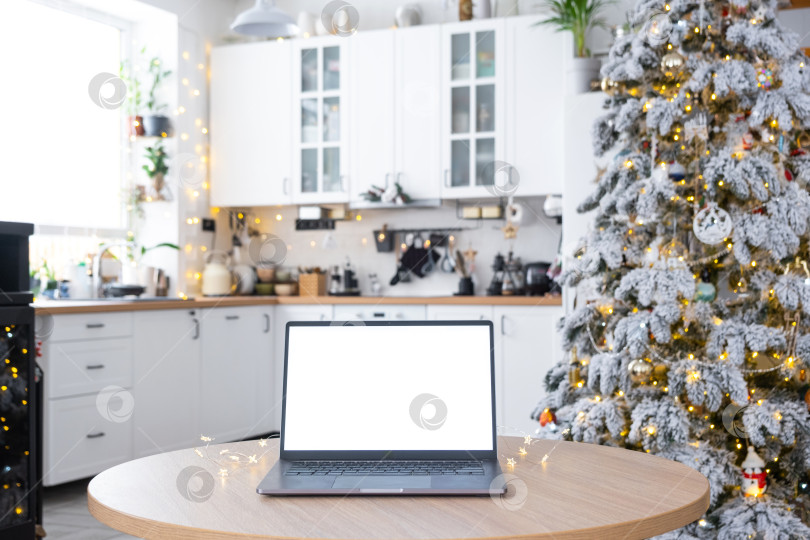 Скачать Макет ноутбука с белым экраном в уютной рождественской кухне, украшенной белыми гирляндами и рождественской елкой. Сезонная удаленная работа, интернет, шопинг, покупка подарков на Рождество и Новый год. фотосток Ozero