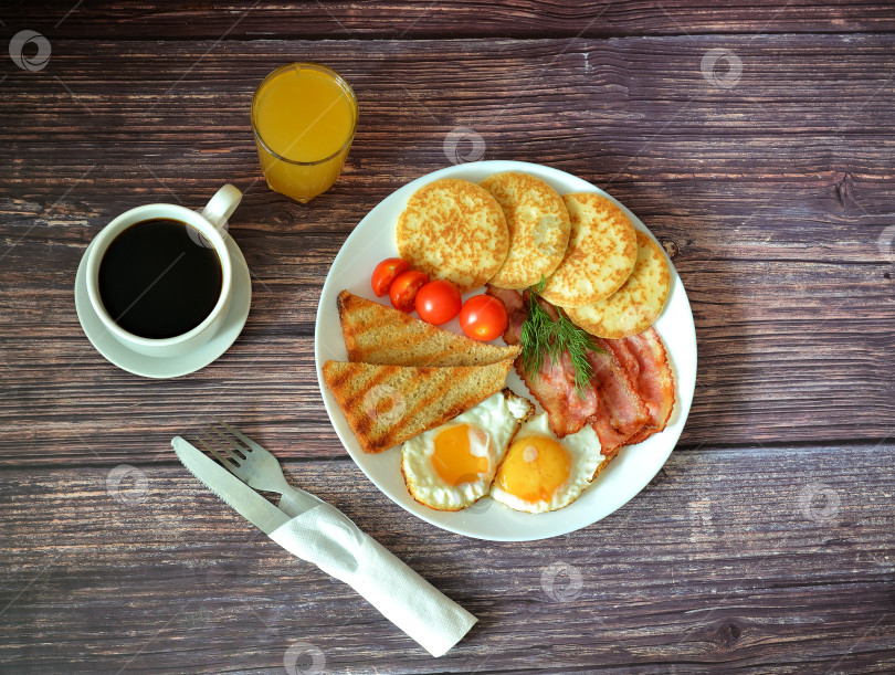 Скачать Завтрак: тарелка с омлетом, гренками, хрустящим беконом, блинчиками и помидорами черри на деревянном столе, салфетка и столовые приборы рядом. фотосток Ozero