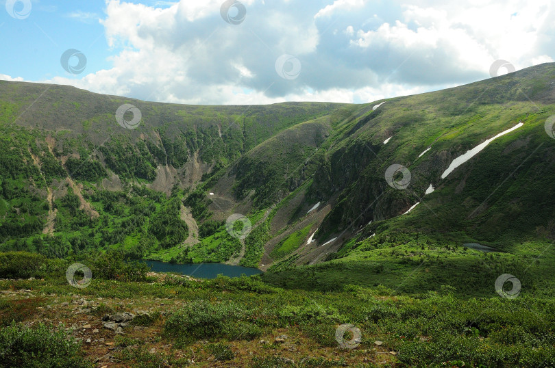 Скачать Вид с вершины холма на небольшое красивое озеро в низинах гор и остатки снега на склонах под летним облачным небом. фотосток Ozero