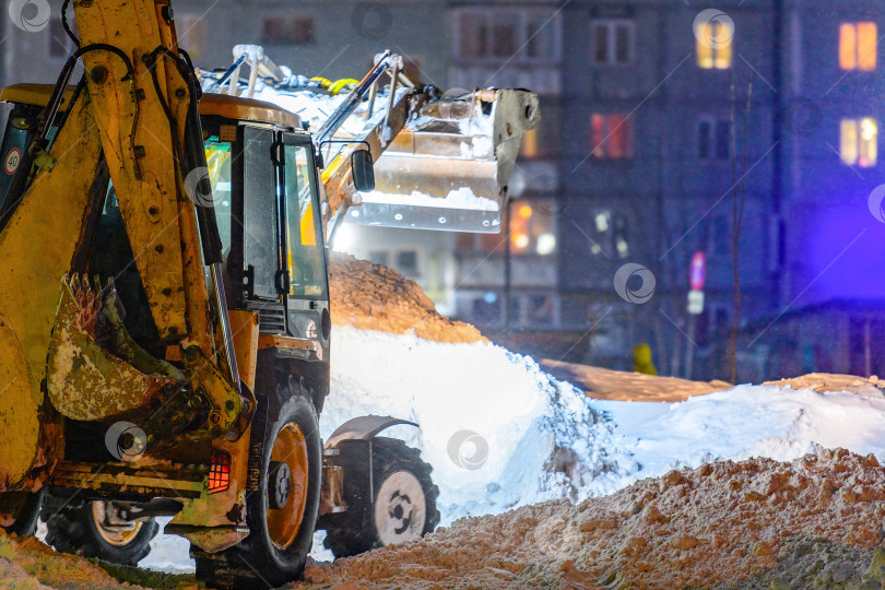 Скачать Снегоуборочная машина, трактор убирают снег на проезжей части улицы в снежную бурю ночью. Служба уборки снега, работающая зимой в городских дворах. фотосток Ozero