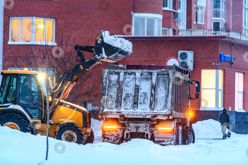 Скачать Расчистка снега коммунальными службами на городских улицах зимой во время метели или снегопада. Снегоуборочный трактор или снегоуборочная машина не в фокусе загружает снег в кузов грузовика. фотосток Ozero