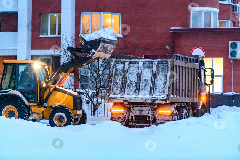 Скачать Расчистка снега коммунальными службами на городских улицах зимой во время метели или снегопада. Снегоуборочный трактор или снегоуборочная машина не в фокусе загружает снег в кузов грузовика фотосток Ozero