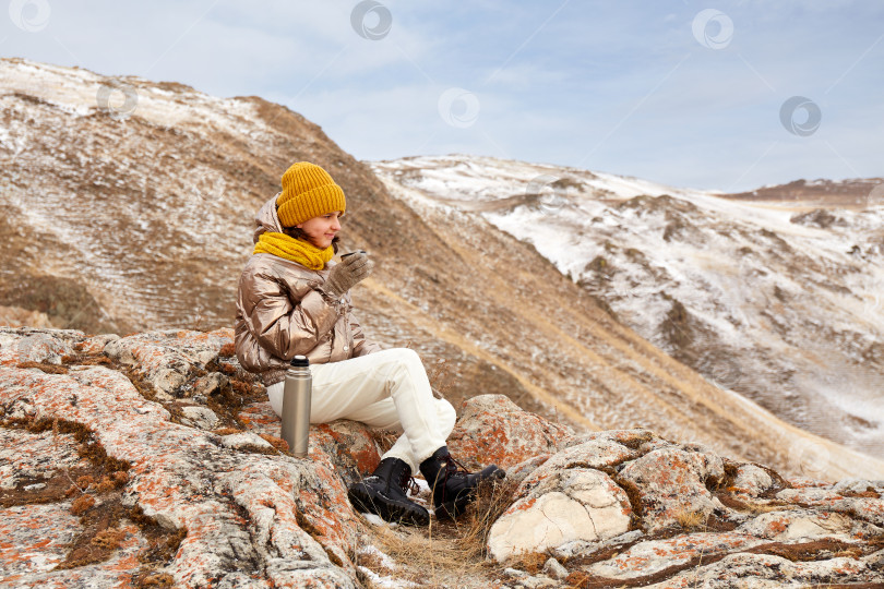 Скачать Молодая красивая девушка-путешественница сидит на камне и держит в руке термос. фотосток Ozero