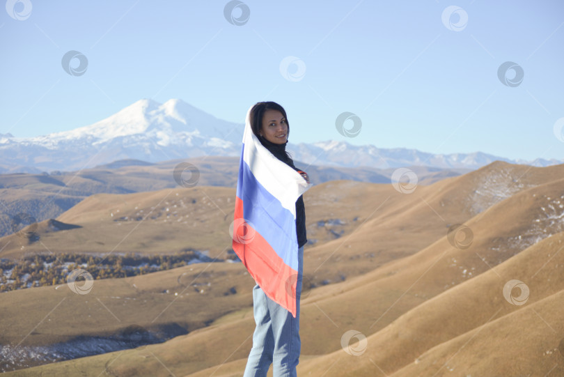 Скачать Молодая брюнетка стоит на фоне заснеженной горы Эльбрус, глядя в камеру, российский флаг покрывает ее плечи. Триколор на фоне заснеженной горы Эльбрус фотосток Ozero