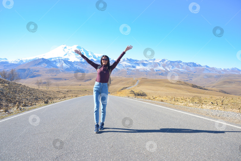Скачать Счастливая путешественница с рюкзаком за спиной и поднятыми вверх руками радуется своей поездке в горы. Эльбрус в ясный осенний день. Вид на Эльбрус, Северный Кавказ, Россия фотосток Ozero