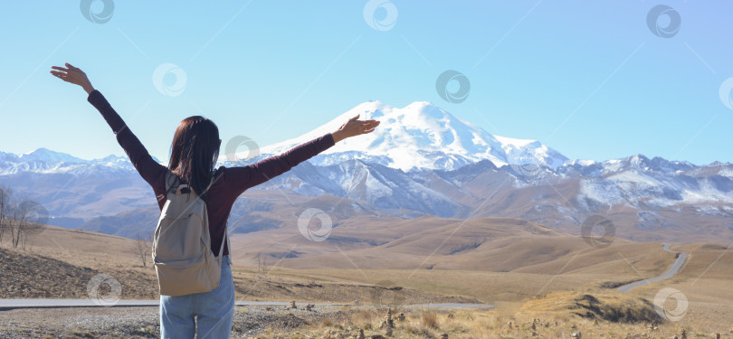 Скачать Счастливая путешественница с рюкзаком за спиной и поднятыми вверх руками любуется заснеженным Эльбрусом в ясный осенний день. Вид на Эльбрус, Северный Кавказ, Россия фотосток Ozero