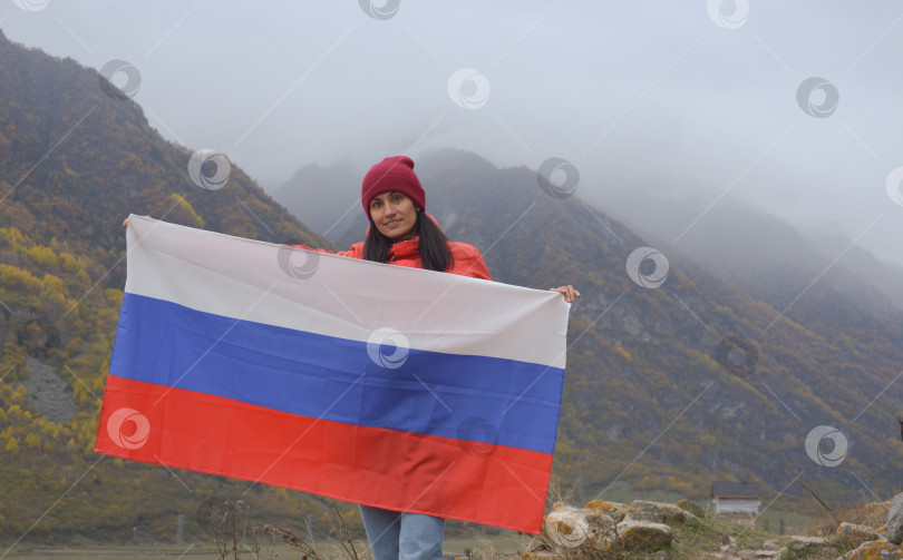Скачать Крупный план российского флага, красиво развевающегося в руках женщины на фоне Кавказских гор в пасмурный туманный день. День независимости России фотосток Ozero