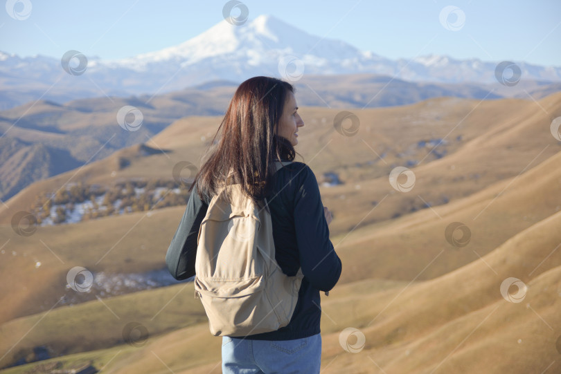Скачать Молодая брюнетка с рюкзаком стоит на фоне заснеженной вершины Эльбруса и смотрит в сторону гор. фотосток Ozero