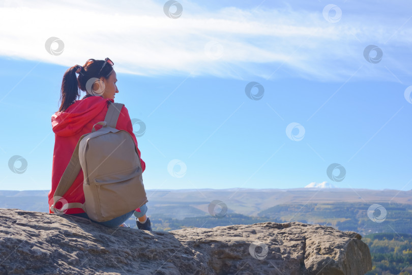 Скачать Девушка-туристка с рюкзаком сидит на вершине горы и наслаждается прекрасными видами. Прогуливающаяся женщина в оранжевой куртке отдыхает на скале, глядя на прекрасный солнечный пейзаж Кавказских гор. фотосток Ozero