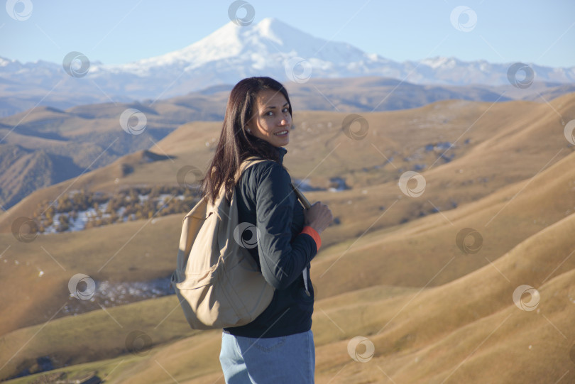 Скачать Молодая брюнетка с рюкзаком стоит на фоне заснеженной горы Эльбрус и смотрит в камеру. фотосток Ozero
