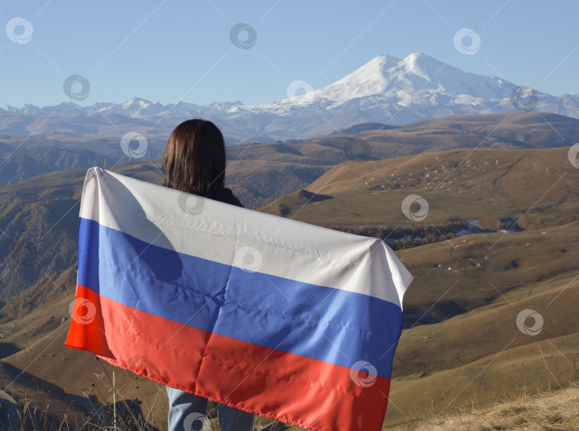 Скачать Молодая брюнетка стоит на фоне заснеженной горы Эльбрус, любуясь красотой гор, держа в руках российский флаг. Триколор на фоне заснеженной горы Эльбрус фотосток Ozero