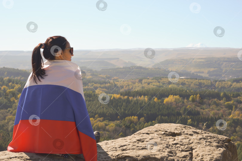 Скачать Девушка-туристка с российским флагом на плечах сидит на вершине горы и наслаждается прекрасным видом на Кавказские горы. фотосток Ozero