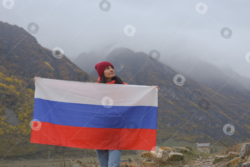 Скачать Крупный план российского флага, красиво развевающегося в руках женщины на фоне Кавказских гор в пасмурный туманный день. День независимости России фотосток Ozero