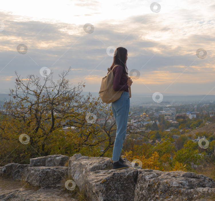 Скачать Девушка с рюкзаком на вершине горы смотрит на город внизу на осеннем закате. Пеший туризм. Природа фотосток Ozero