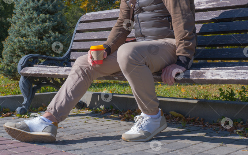 Скачать Молодой человек сидит на скамейке в осеннем парке, пьет кофе, отдыхает, расслабляется. Концепция городского образа жизни. фотосток Ozero