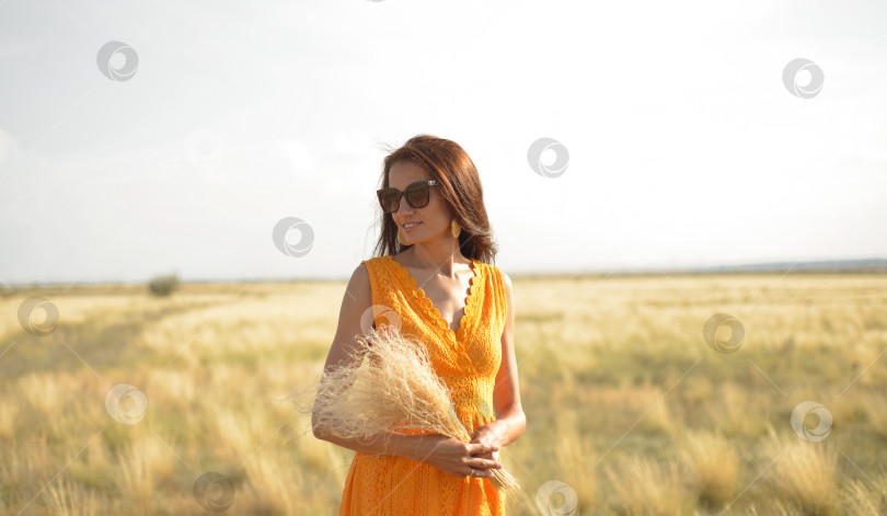 Скачать Красивая девушка-брюнетка в оранжевом платье и солнцезащитных очках с букетом полевой травы наслаждается природой летом. фотосток Ozero