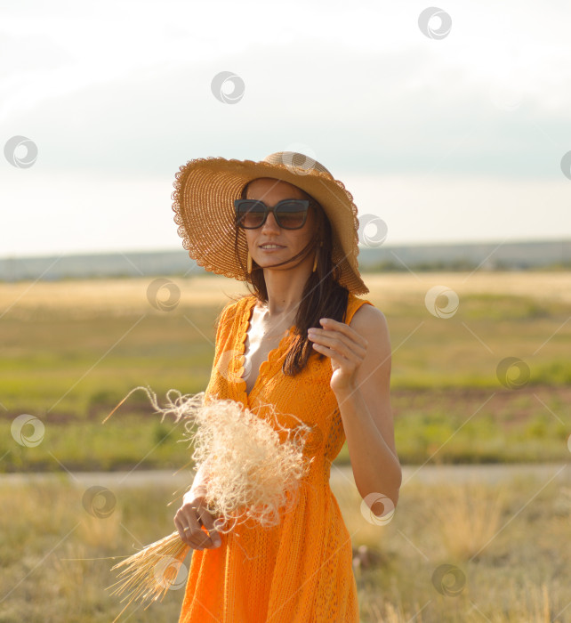 Скачать Молодая женщина в оранжевом платье и соломенной шляпе стоит на поле в лучах заходящего солнца фотосток Ozero