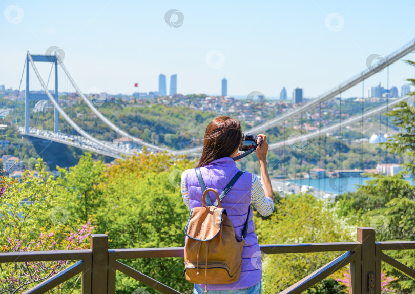 Скачать Молодая туристка с рюкзаком фотографирует городской пейзаж с панорамным видом на мост Фатиха Султана Мехмета. Вид сзади. Парк Отагтепе фотосток Ozero