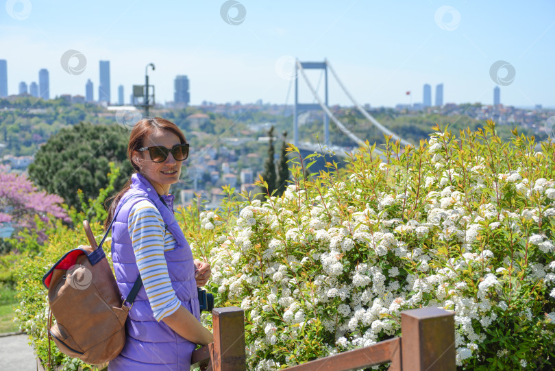 Скачать Молодая красивая туристка с рюкзаком смотрит в камеру на фоне городского пейзажа с видом на мост Фатиха Султана Мехмета. Вид сзади. Парк Отагтепе фотосток Ozero