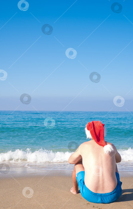 Скачать Мужчина в шляпе Санта-Клауса сидит на берегу моря, вид сзади. Рождественский Санта-Клаус отдыхает на песке тропического пляжа в океане - направления для новогодних путешествий, отдых в жарких странах, концепция скидок на поездки фотосток Ozero