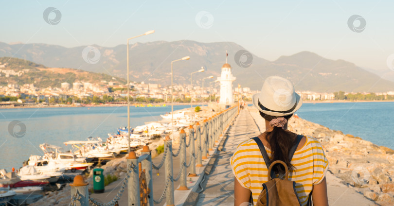 Скачать Девушка-туристка в шляпе с рюкзаком на фоне набережной с маяком в жаркий летний день, вид сзади. фотосток Ozero
