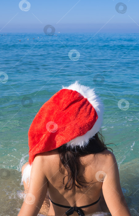 Скачать Фотография молодой женщины с длинными волосами и в шапке Санта-Клауса, смотрящей на море. Концепция путешествий и туризма на Рождество, Новый год и зимние каникулы. фотосток Ozero