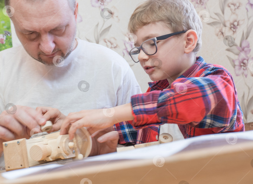 Скачать Концепция счастливого дня отца и детства. Отец и сын-мальчик в очках работают с ручными инструментами, используя отвертку, собирая деревянный конструктор дома за столом. фотосток Ozero
