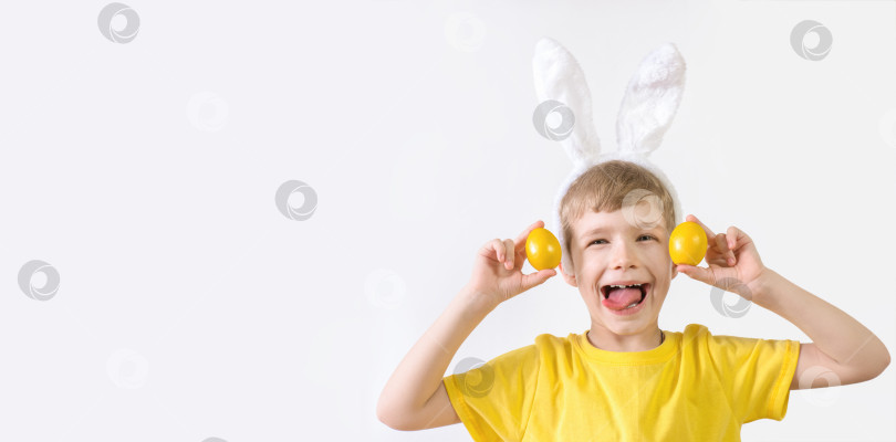 Скачать Счастливый ребенок показывает язык в желтой футболке с заячьими ушками и разноцветными яйцами на белом фоне. Концепция счастливой Пасхи. фотосток Ozero