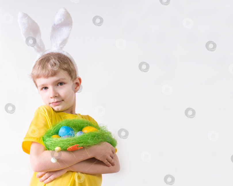 Скачать Улыбающийся счастливый ребенок в желтой футболке с заячьими ушками и с разноцветными яйцами в корзинке на белом фоне. Концепция счастливой Пасхи с местом для текста. фотосток Ozero