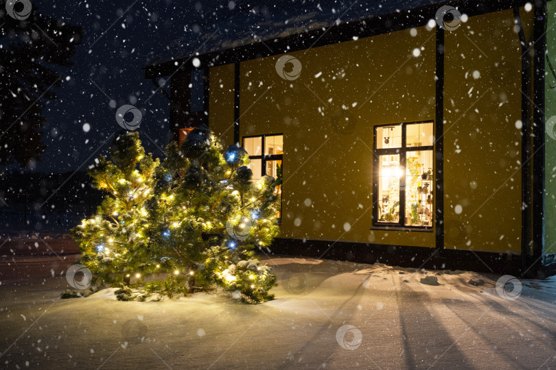 Скачать Уютное праздничное окно дома снаружи с теплым светом гирлянд fairy lights внутри - празднуйте Рождество и Новый год в теплом доме. Рождественская елка, боке, снег на соснах и снегопад фотосток Ozero
