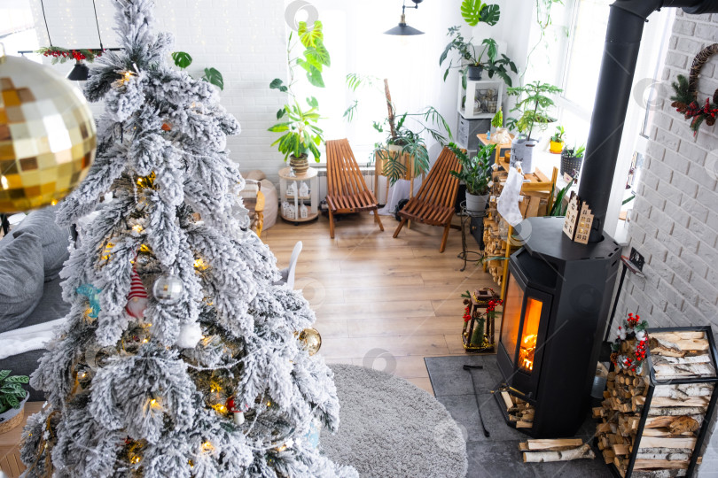 Скачать Праздничный белый современный интерьер дома оформлен к Рождеству и Новому году в стиле лофт с черной печью, камином, рождественской елкой. Растения в горшках, дрова в поленнице фотосток Ozero